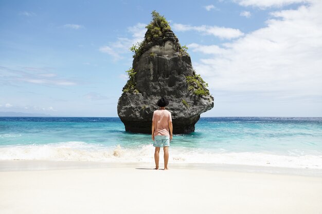 Voyage, aventure et tourisme. Homme aux pieds nus à la mode, vêtu d'un short, d'un t-shirt et d'un chapeau méditant au bord de la mer, debout devant l'île de pierre. Élégant touriste caucasien admirant la belle vue