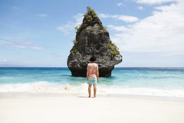 Voyage, aventure et tourisme. Homme aux pieds nus à la mode, vêtu d'un short, d'un t-shirt et d'un chapeau méditant au bord de la mer, debout devant l'île de pierre. Élégant touriste caucasien admirant la belle vue