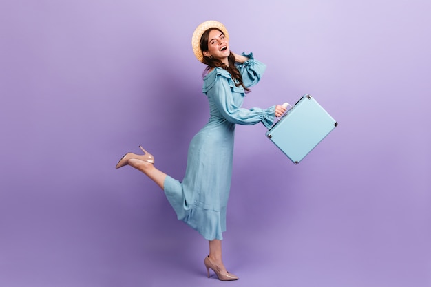 Un voyageur élégant s'exécute sur un mur lilas. Le modèle en chapeau et talons garde la sacoche.