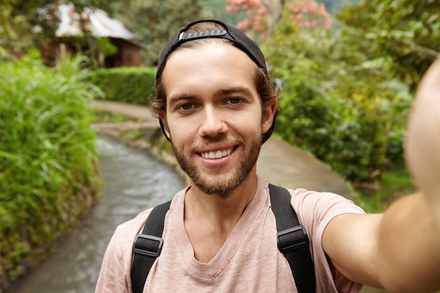 Photo gratuite voyageur à la mode attrayant avec barbe portant un sac à dos prenant selfie, posant sur une route de campagne. voyage, aventure