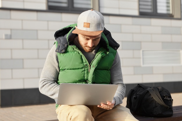 Voyageur à la mode jeune homme assis sur un banc avec sac, tenant un ordinateur portable sur ses genoux, réservation d'un appartement tout en restant dans une ville étrangère en voyage d'affaires. Technologie, voyages et style de vie