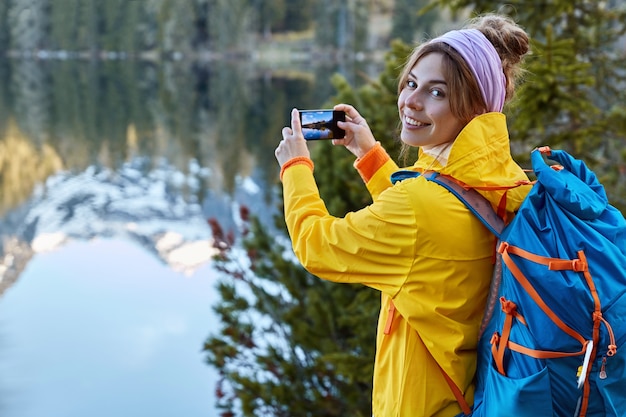 Le voyageur touristique tient un téléphone intelligent dans les mains, fait une photo de paysage panoramique en voyage, admire le voyage dans les montagnes, pose près du lac