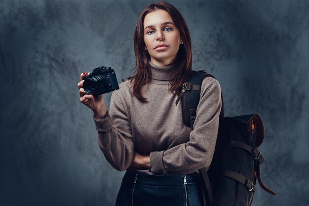 Une voyageuse brune avec sac à dos tient un appareil photo compact.