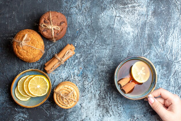 Vue aérienne de citrons frais et main tenant une tasse de thé noir à la cannelle divers biscuits empilés sur fond sombre