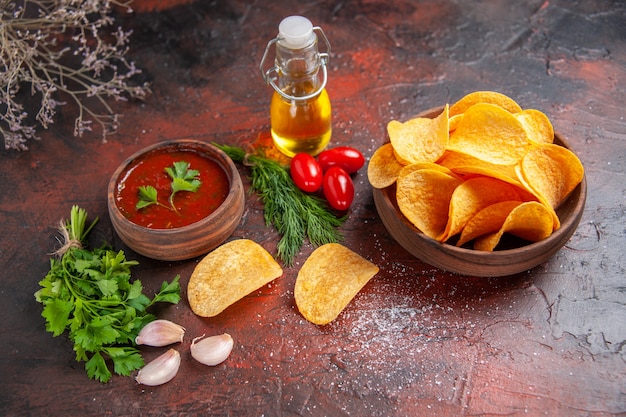 Vue aérienne de délicieuses chips croustillantes de pommes de terre faites maison dans un petit bol marron bouteille d'huile tomates vertes ketchup à l'ail et poivre haché sur table sombre