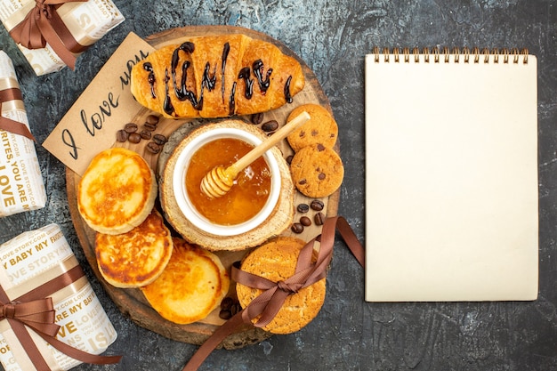 Vue aérienne d'un délicieux petit-déjeuner avec des crêpes aux croissants, des biscuits empilés au miel, un beau cadeau pour un bien-aimé et un nootbook en spirale sur fond sombre