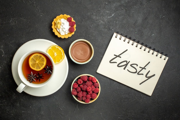 Vue aérienne du cahier avec inscription savoureuse et une tasse de thé noir au citron servie avec du miel de framboise au chocolat sur fond sombre