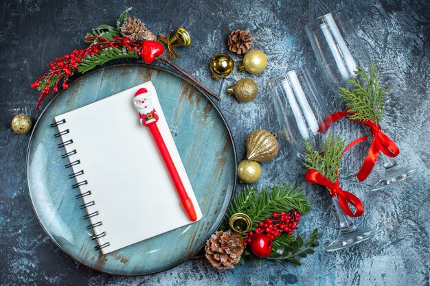 Vue aérienne de gobelets en verre avec cahier à ruban rouge avec stylo sur une plaque bleue et accessoires de décoration de branches de sapin sur fond sombre