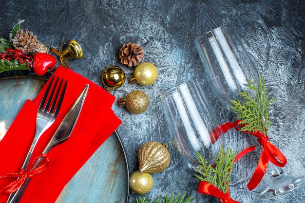 Vue aérienne de gobelets en verre avec des couverts en ruban rouge sur une assiette bleue et des accessoires de décoration de branches de sapin sur fond sombre