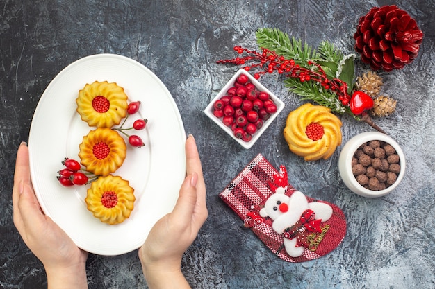 Vue aérienne de la main tenant une assiette blanche avec de délicieux biscuits accessoire de décoration chaussette du père noël et cornell dans un bol de branches de sapin sur une surface sombre