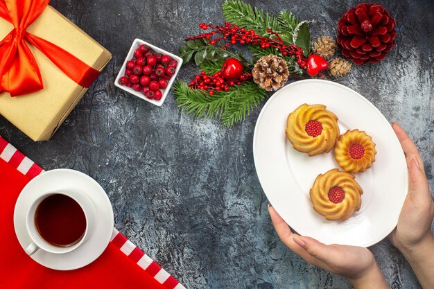 Vue aérienne d'une tasse de thé noir sur une serviette rouge et main tenant des biscuits sur une assiette blanche cadeau d'accessoires du nouvel an avec ruban rouge sur une surface sombre