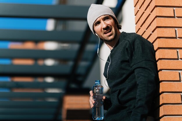 Vue en angle bas d'un athlète masculin fatigué prenant une pause après avoir fait de l'exercice et buvant de l'eau tout en s'appuyant sur un mur