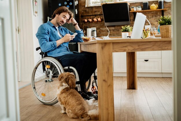 Vue en angle bas d'un entrepreneur souriant dans la messagerie texte en fauteuil roulant sur un téléphone mobile tout en prenant sa pause déjeuner à la maison