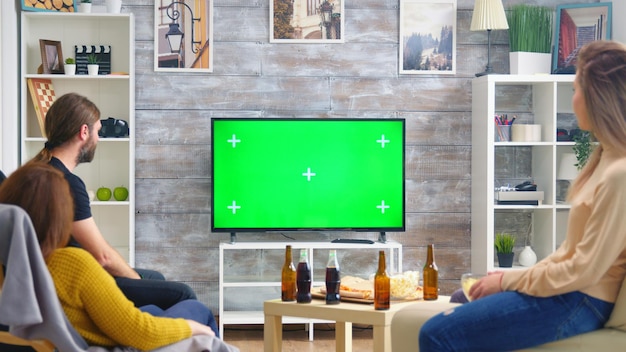 Vue arrière d'amis buvant de la bière et regardant des sports à la télévision avec écran vert dans le salon.