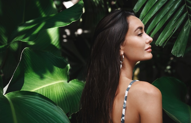 Photo gratuite vue arrière de la belle femme brune avec bronzage doré, vêtu d'un bikini, debout de profil dans les feuilles vertes.