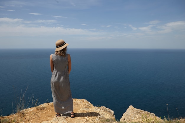 Photo gratuite vue arrière de la belle femme portant un chapeau de paille et une robe maxi d'été debout au sommet de la montagne et contemplant une magnifique mer sans fin devant elle. concept de vacances, de voyages et de bord de mer