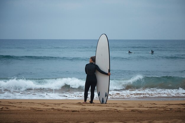 Vue arrière sur la belle jeune fille de surf étreignant son longboard au bord de l'océan et regardant les vagues avant de surfer