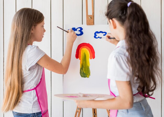 Vue arrière de deux filles peignant les nuages sur une toile avec un pinceau