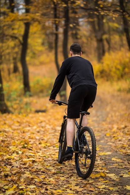 Vue arrière du jeune bel homme monté sur un vélo sur la route forestière parmi les arbres au coucher du soleil. Sport et mode de vie sain. Voyage dans la forêt tropicale