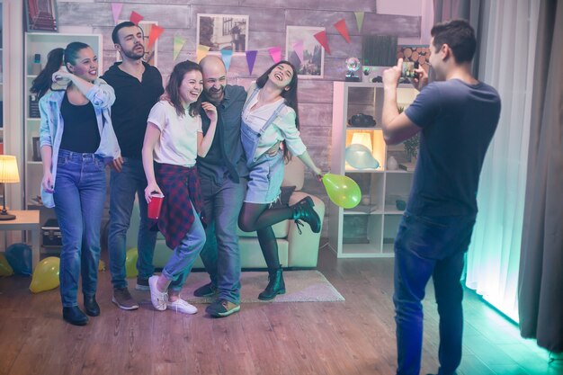 Vue arrière du jeune homme heureux de prendre des photos d'amis proches lors d'une fête dans un appartement.