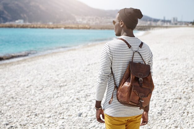 Vue arrière du routard africain face à la mer dans des vêtements à la mode, voyageant seul dans une station balnéaire européenne, admirant l'eau bleu ciel et les montagnes, pensant à quelque chose de secret et d'intime