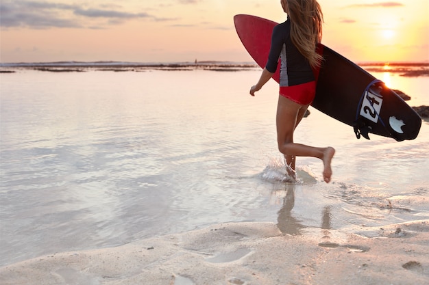 Photo gratuite vue arrière du surfeur fille en maillot de bain, porte planche sous le bras, prêt à conquérir une vague géante, se jette dans l'océan