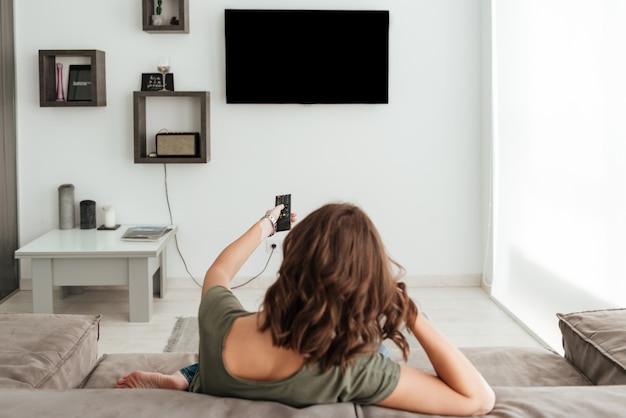 Photo gratuite vue arrière d'une femme décontractée assise sur un canapé et regarder la télévision à la maison