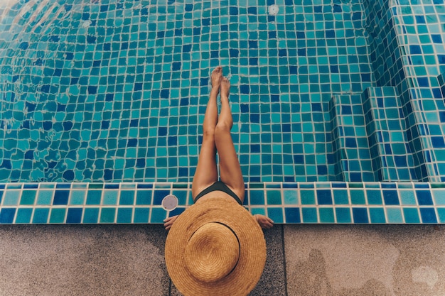 Photo gratuite vue arrière d'une femme gracieuse en maillot de bain et chapeau assis près de la piscine