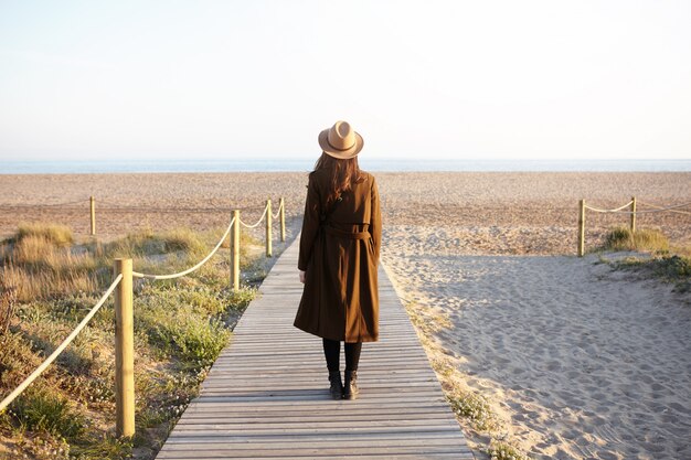 Vue arrière de la femme à la mode avec des cheveux noirs lâches debout seul sur la promenade en direction de la mer. Une jeune femme méconnaissable en chapeau et manteau est venue dans l'océan pour se vider la tête tout en faisant face au stress au travail