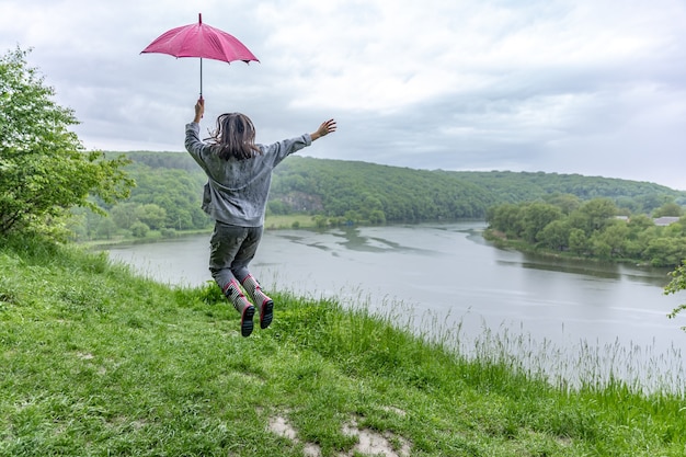 Vue arrière d'une fille sous un parapluie sautant près d'un lac dans une zone montagneuse par temps de pluie.