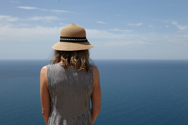 Vue arrière gros plan d'une femme blonde élancée méconnaissable en robe et chapeau de paille bénéficiant d'un paysage marin incroyable au point de vue. Femme romantique admirant des paysages pittoresques sur un vaste océan calme et un ciel bleu