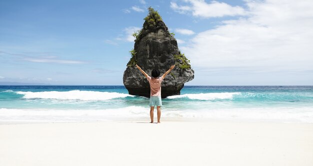 Vue arrière de l'homme en face de l'île rocheuse avec une végétation tropicale, admirant une vue magnifique, debout sur la plage avec l'eau de l'océan azur et le ciel bleu à l'horizon