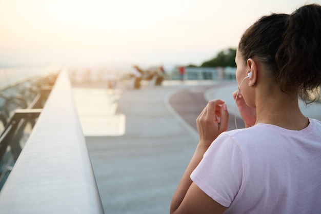 Vue arrière d'une jeune athlète en t-shirt rose mettant des écouteurs et se préparant pour le jogging matinal, regardant le beau lever de soleil au bout du pont tôt le matin