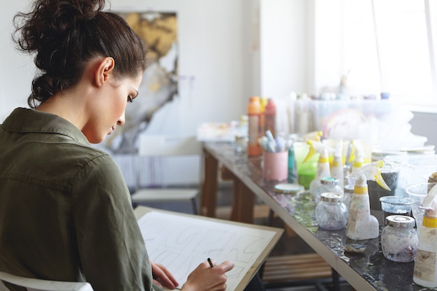 Vue arrière de la jeune créatrice européenne sérieuse et concentrée aux cheveux noirs travaillant sur une nouvelle collection de bijoux ou de vêtements dans son atelier spacieux et lumineux, inspirée. Processus de création