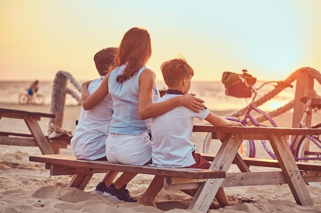 Vue arrière d'une jolie famille - mère avec ses fils assis sur un banc et regarde le coucher du soleil sur le littoral.