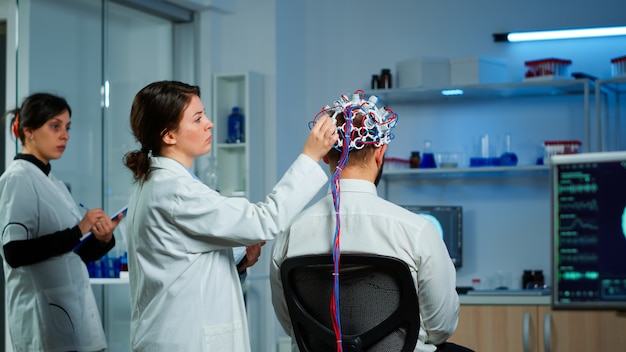 Vue arrière d'un patient portant un casque à balayage d'ondes cérébrales performant assis dans un laboratoire de recherche neurologique pendant qu'un chercheur médical l'ajuste, examinant la saisie du système nerveux sur une tablette.