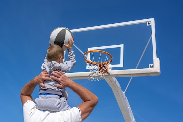 Photo gratuite vue arrière d'un petit-fils assis sur l'épaule de son grand-père jetant un ballon de basket dans un cerceau