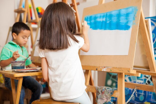 Vue arrière d'une petite fille peignant le ciel d'un paysage pour son cours d'art