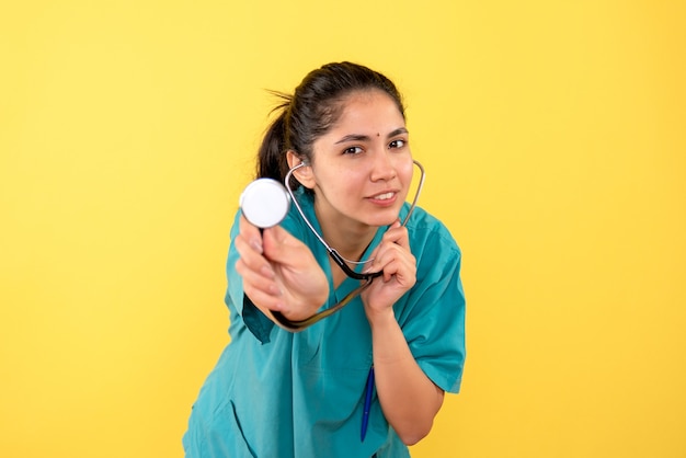 Vue avant béate femme médecin en uniforme montrant un stéthoscope sur fond jaune