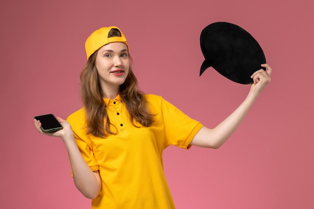 Vue avant de courrier féminin en uniforme jaune et cape tenant une pancarte noire et un smartphone sur un mur rose en uniforme de livraison d'emploi