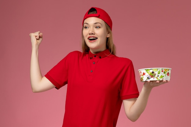 Vue Avant De Courrier Féminin En Uniforme Rouge Et Cape Tenant Le Bol De Livraison Et Acclamant Le Mur Rose, La Livraison D'uniforme De Service