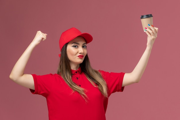 Vue avant de courrier féminin en uniforme rouge et cape tenant la tasse de café de livraison et fléchissant sur l'uniforme de travailleur de livraison de travail de fond rose