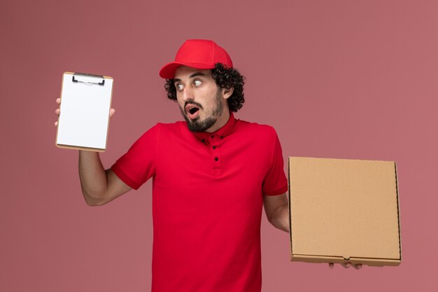 Vue avant de courrier masculin en chemise rouge et cape tenant la boîte de nourriture de livraison et le bloc-notes sur le mur rose