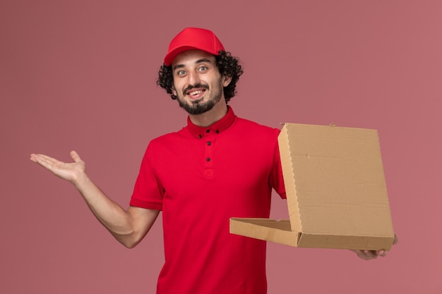 Vue avant de courrier masculin en chemise rouge et cape tenant la boîte de nourriture de livraison vide souriant sur le mur rose clair