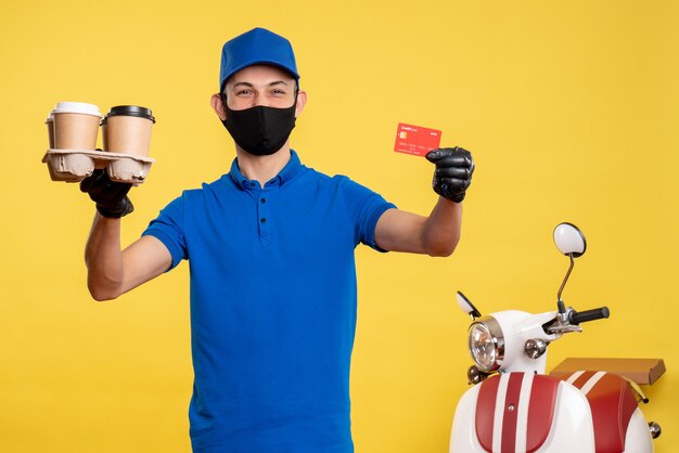Vue avant de courrier masculin en masque tenant le café et la carte bancaire sur l'uniforme de travail jaune covid- livraison service d'emploi pandémique
