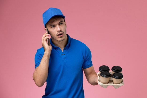 Vue avant de courrier masculin en uniforme bleu parlant au téléphone tenant du café sur le mur rose, la prestation de services uniforme des travailleurs