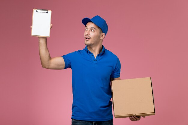 Vue avant de courrier masculin en uniforme bleu tenant le bloc-notes avec boîte de nourriture sur le mur rose clair, la prestation de services uniforme des travailleurs