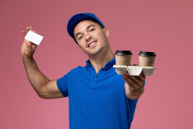 Vue avant de courrier masculin en uniforme bleu tenant des tasses à café carte blanche sur mur rose, prestation de services uniforme de travailleur d'emploi
