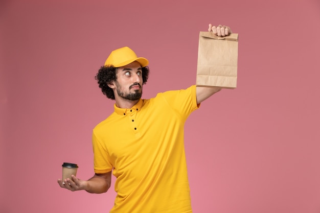 Vue Avant De Courrier Masculin En Uniforme Jaune Et Cape Tenant La Tasse De Café De Livraison Et Le Paquet De Nourriture Sur Le Bureau Rose Uniforme De Travail De L'entreprise De Service D'emploi