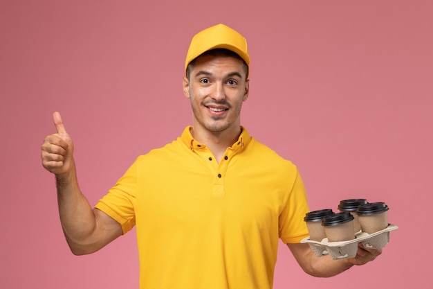 Vue avant de courrier masculin en uniforme jaune souriant et tenant des tasses de café de livraison sur le bureau rose